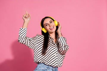 Joyful young woman in striped shirt and yellow headphones dances against pink background, expressing happiness and fun
