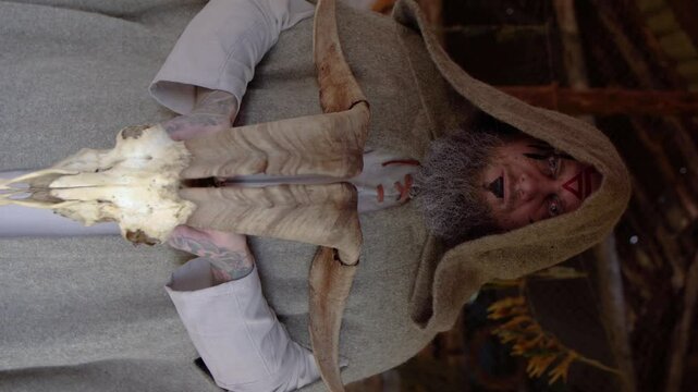 Portrait of a bearded man with face paint in a hood holding an animal skull with horns looking at camera. 4K