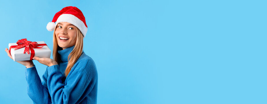 Happy lady in sweater and Santa hat, holding gift box and looking back at free space, isolated on blue studio background, panorama. Great gift idea for New Year, Christmas celebration
