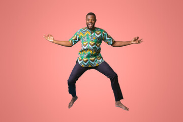 Carefree black man in african costume posing while jumping up on blue studio background. Smiling african american guy wearing traditional clothes, full size photo. Traditions and culture concept