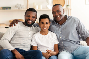 Obraz premium Portrait of smiling millennial black dad, preteen son and grandfather posing to camera at home, sitting on couch in living room, smiling