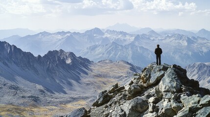 A lone figure stands atop a rocky peak, gazing at the vast expanse of rugged mountain ranges under a serene, cloud-streaked sky.