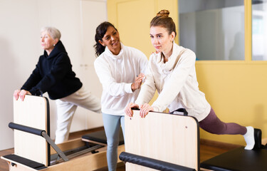 Two active women practicing Pilates in a fitness studio perform an exercise using a reformer bed, where a female instructor ..helps them do it correctly