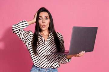 Young brunette woman in striped shirt holding laptop with surprise on pink background