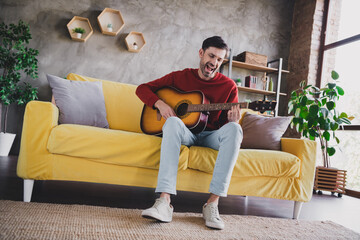 Young man enjoying his day off playing guitar on a cozy yellow sofa in stylish apartment filled with natural light and autumn decor