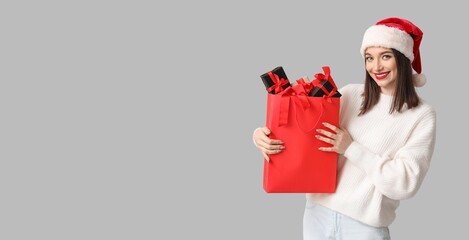 Pretty young woman in Santa hat holding shopping bag full of Christmas gifts on grey background with space for text