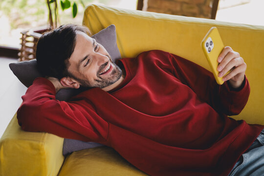 Young man relaxing at home on comfy sofa, enjoying leisure time with smartphone in hand, wearing casual red pullover.
