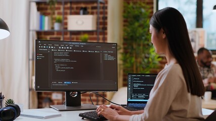 Asian woman is seen typing code for an app development project on her computer, working from a home office. IT specialist engineer focuses on software programming and remote tech work. Camera B.