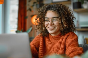 Smiling woman working from home on laptop wearing glasses and orange sweater, curly hair, studying online, watching webinar, remote learning, virtual meeting, freelance, casual attire