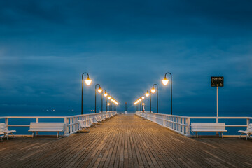 Wooden pier illuminated by lampposts at dusk with white benches on each side and calm baltic sea in the background, located in sopot, poland
