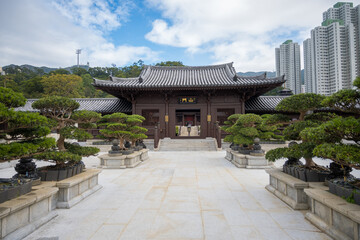 Chi Lin Nunnery entrance with a brown wooden structure and a tiled roof, flanked by rows of bonsai trees in stone planters. The path leading to the temple is paved with gray stone tiles.