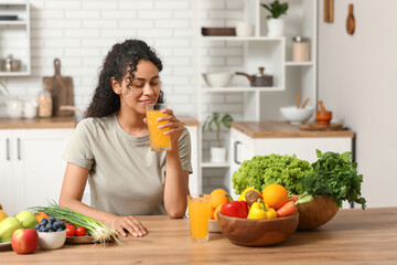 Young happy African-American woman with glasses of juice, different vegetables and fruits sitting in kitchen. Independence from Meat Day