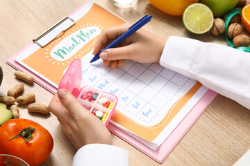 Female nutritionist writing diet plan and holding pills near healthy food on table