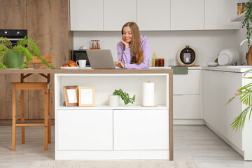 Young woman using modern laptop at table in kitchen