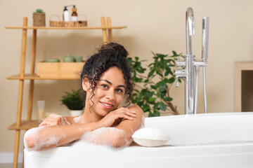 Beautiful young happy African-American woman taking bath with sponge and soap foam at home