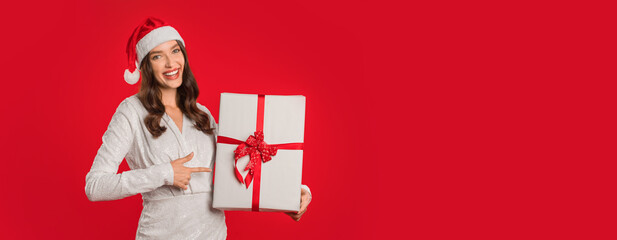 Joyful woman celebrates Christmas season pointing at wrapped gift box with red bow, posing in elegant white dress and Santa hat on studio backdrop. Winter holiday promotions and New Year sale