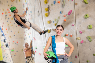 Happy sporty young female holding helmet and blue ropes while standing against concrete wall background. Rock, mountain, industrial albinism climbing concept