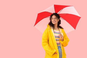 Happy young African-American woman in raincoat with umbrella on pink background