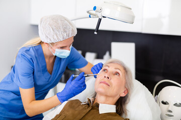 Fototapeta premium Female cosmetologist in mask and medical gloves making injections of botulotoxin in face of elderly woman.