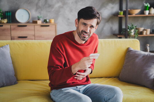 Young man with a stomach ache sitting on a yellow sofa indoors wearing a red casual pullover, looking uncomfortable as he holds medication.