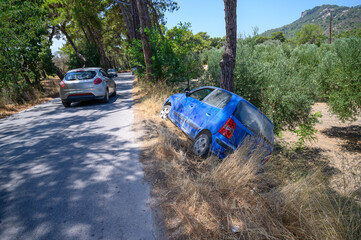 Crashed blue car out of street next to tree – back view, other cars on street