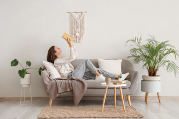 Fototapeta premium Pretty young woman reading book on sofa near white wall