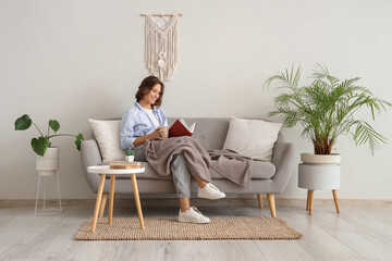 Fototapeta premium Pretty young woman with cup of tea reading book on sofa at home