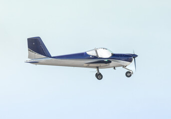 Down the wingtip, looking broadside at an airplane as it flies by. Blue and gray light aircraft powered by a propeller. Fixed landing gear aircraft built from a kit, large glass canopy