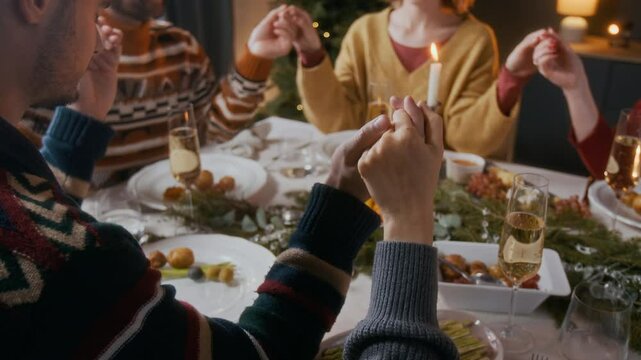 Close up view of family members sitting around festive dinner table, holding hands and saying prayer before Christmas meal during traditional home celebration