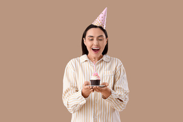 Young woman in striped pajamas with party hat and Birthday cupcake on beige background