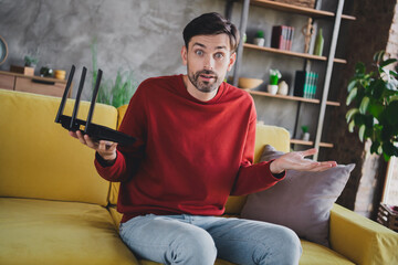 Young man with puzzled expression holding router on yellow sofa in modern apartment