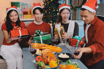 Group of young Asian man and women as friends having fun at a New Year's celebration, holding gift boxes standing by Christmas tree decoration, midnight countdown Party at home with holiday season.