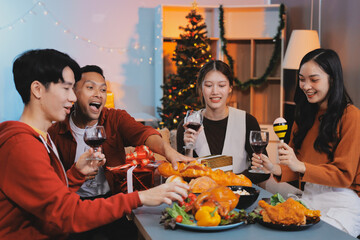 Group of young Asian man and women as friends having fun at a New Year's celebration, holding gift boxes standing by Christmas tree decoration, midnight countdown Party at home with holiday season.