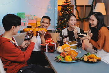 Group of young Asian man and women as friends having fun at a New Year's celebration, holding gift boxes standing by Christmas tree decoration, midnight countdown Party at home with holiday season.