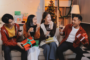 Group of young Asian man and women as friends having fun at a New Year's celebration, holding gift boxes standing by Christmas tree decoration, midnight countdown Party at home with holiday season.