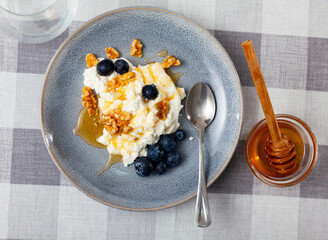 Fresh farm cottage cheese filled with large amount of liquid summer honey is served on plate, decorated with walnut and blueberry