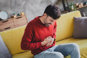 Young man experiencing stomach pain while seated on a sofa at home, in casual jumper, a moment of...