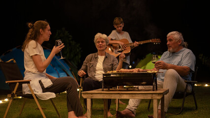 Family barbecue during a camping trip is grilling food on a portable barbecue , with holding glasses of wine ,play guitar as they enjoy the moment together shared moments family holiday camping trip.