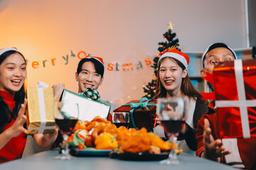 Group of young Asian man and women as friends having fun at a New Year's celebration, holding gift boxes standing by Christmas tree decoration, midnight countdown Party at home with holiday season.
