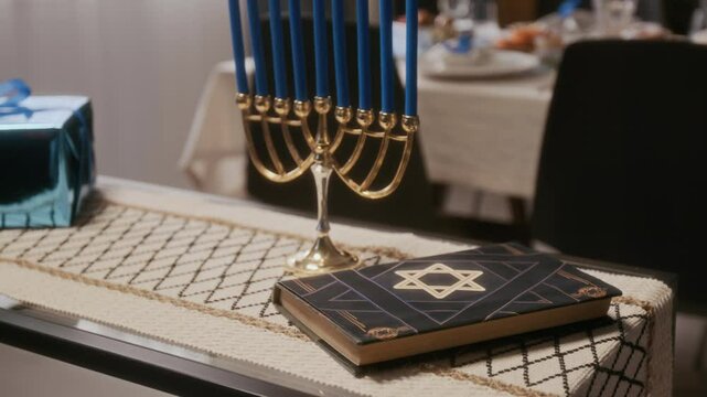 Close up view of Hanukkah menorah with blue candles, Hebrew Bible and gift box placed on side table in living room prepared for holiday dinner