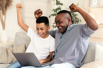 Fototapeta premium Joyful black granddad and grandson watching sports on laptop at home and emotionally cheering, raising fists in excitement, free space