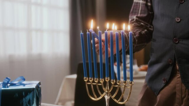 Close up view of unrecognizable man lighting blue candles on Hanukkah menorah with wrapped gift nearby