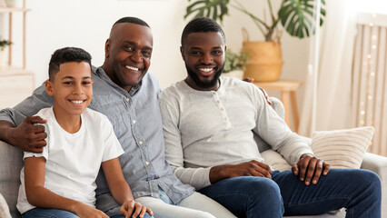 Africam American Grandfather With Son And Grandson Relaxing Together At Home, Embracing On Sofa, Looking At Camera And Smiling