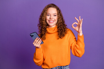 Portrait of pretty young girl smart phone okey symbol wear orange sweater isolated on purple color background