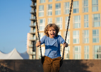 Kid swinging on chain swing on city kids playground. Swing ride. Cute child having fun on a swing on summer sky background. Blonde little boy swings at kid playground. Child swinging high.