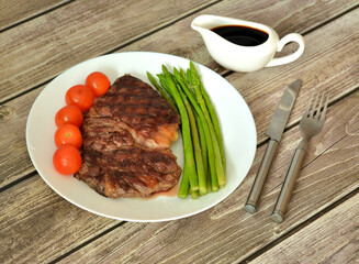 Plate with freshly fried beef steak, cherry tomatoes and asparagus on a wooden table, rows of cutlery and gravy boat.