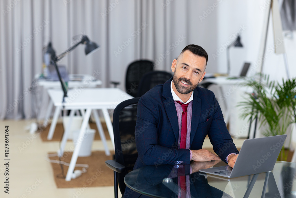 Poster a man in a suit is seated at a glass desk in a contemporary office. he smiles while using a laptop, 