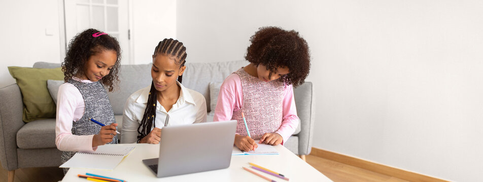 African American mother assists her two daughters with their homework in a comfortable living room, surrounded by colorful stationary and a laptop on the table as they focus on their tasks.