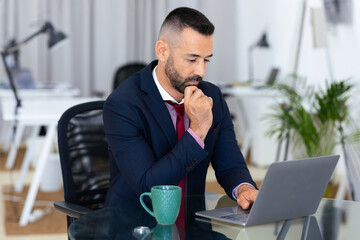 A man in a suit is focused on his laptop in a contemporary office. He appears to be deep in...