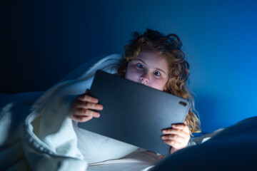 Kid lying on bed and surfing Internet on tablet in dark room. Boy reading Tablet in bed at night. Kid in pajamas using tablet before going to bed. Kid using tablet gadgets.